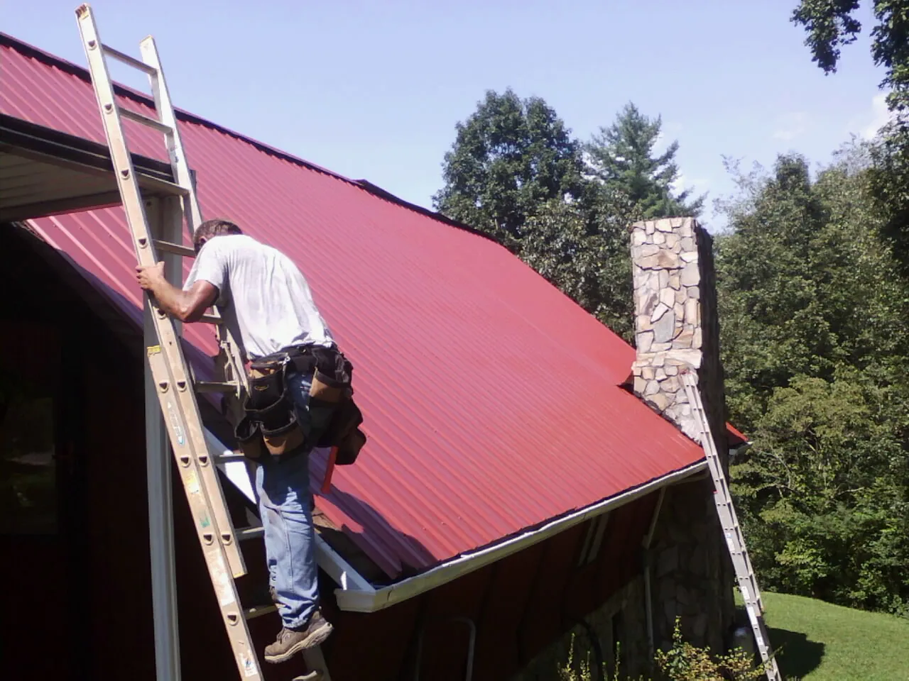 Roofer on a ladder beside a red metal roof and stone chimney — Soffit & Fascia Repair by Roofing Brothers in Robstown